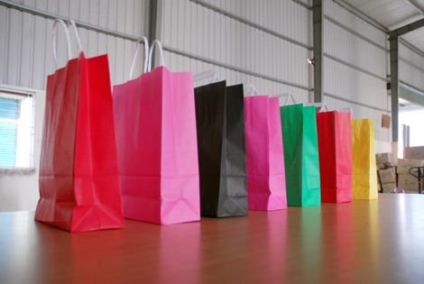 A row of colorful paper shopping bags- red, pink, black, another shade of pink, green, another red, and yellow, standing in a line on a wooden table inside a warehouse setting.
