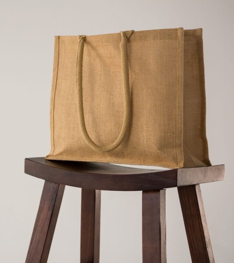 A jute burlap tote bag with thick rope handles sits on a dark wooden stool against a neutral white background.